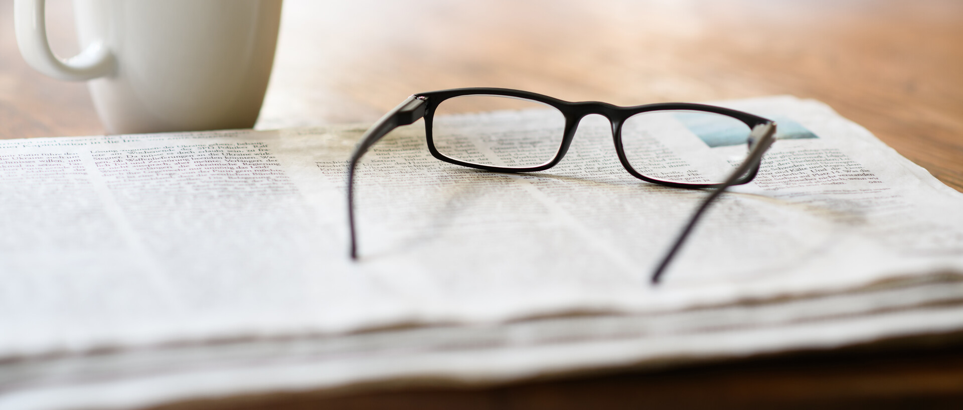 A newspaper with a pair of reading glasses on top. Next to it is a coffee cup