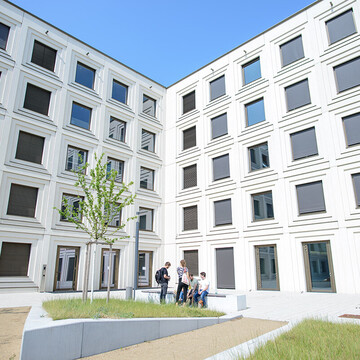 Innenhof des mit zahlreichen Fenstern gebauten Universitätsgebäudes B6 mit kleinen Grünflächen und im Hintergrund sitzen Studierende auf einer Bank.