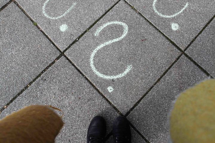 Boden auf dem vier Fragezeichen mit weißer Kreide gemalt wurden. Am unteren Bildrand schwarze Stiefel und schwarze Hose der Kamerafrau zu sehen.