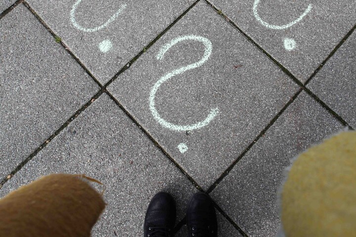 Boden auf dem vier Fragezeichen mit weißer Kreide gemalt wurden. Am unteren Bildrand schwarze Stiefel und schwarze Hose der Kamerafrau zu sehen.
