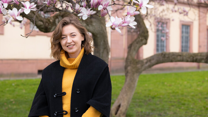 Eine Person trägt einen gelben Rollkragenpullover sowie einen schwarzen Mantel und steht vor einem Magnolienbaum. Die Person heißt Alina Ivanenko.