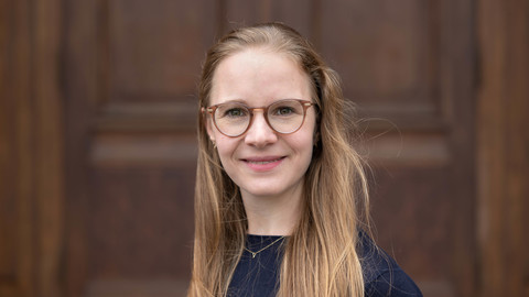 Laura Montenbruck. She is wearing a black sweater and is standing in front of a brown door.