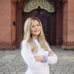 Nadine Wendroswski trägt eine weiße Bluse und hat ihre langen blonden Haare offen. Sie hat die Arme verschränkt und steht auf dem Ehrenhof der Uni Mannheim.