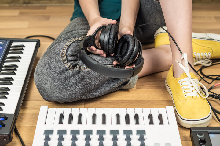 Person mit grauer Jeanshose, gelben Schuhen und Kopfhörer in der Hand sitzt auf dem Boden und gegenüber von zwei auf dem Boden liegenden Pianos.
