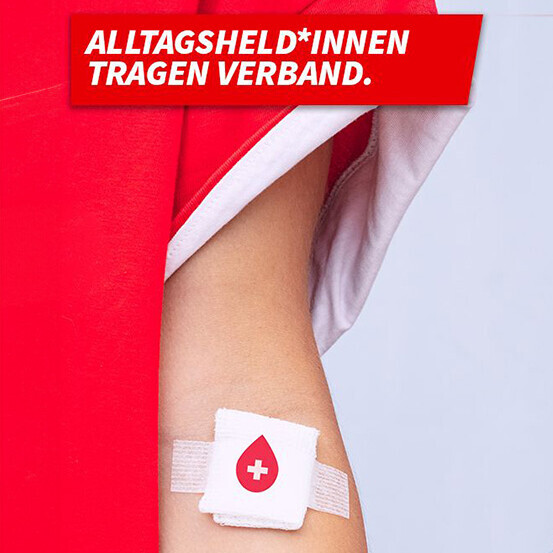 Ein Arm mit einem roten Ärmel und ein Arm mit einem weißen Ärmel heben sich vor einem hellgrauen Hintergrund ab. In den Armbeugen sind zwei Pflaster mit roten Kreuzen zu sehen, die typischerweise nach einer Blutspende aufgeklebt werden. Über dem Bild steht in großer, roter Schrift: „ALLTAGSHELD*INNEN TRAGEN VERBAND.“