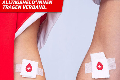 Ein Arm mit einem roten Ärmel und ein Arm mit einem weißen Ärmel heben sich vor einem hellgrauen Hintergrund ab. In den Armbeugen sind zwei Pflaster mit roten Kreuzen zu sehen, die typischerweise nach einer Blutspende aufgeklebt werden. Über dem Bild steht in großer, roter Schrift: „ALLTAGSHELD*INNEN TRAGEN VERBAND.“