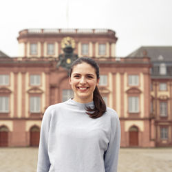 Eine junge Frau steht vor dem Mannheimer Schloss. Sie trägt einen grauen Pullover und einen Pferdeschwanz.