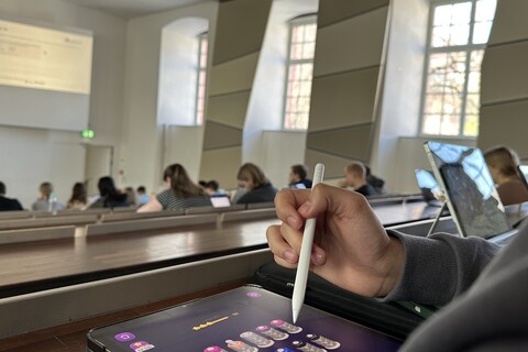 Eine Person sitzt in einer Vorlesung und spielt auf einem Tablet ein Mobilspiel.