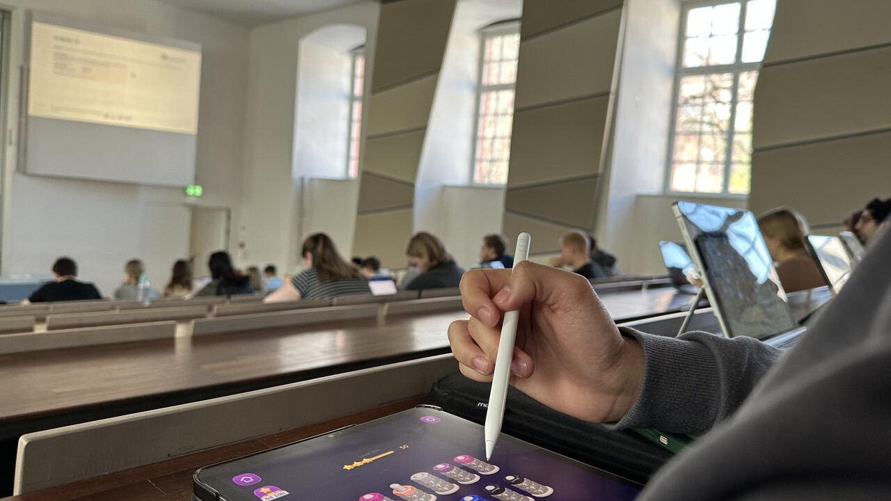 Eine Person sitzt in einer Vorlesung und spielt auf einem Tablet ein Mobilspiel.