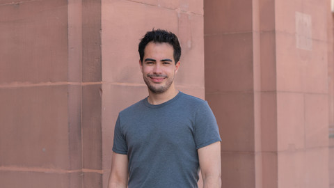 Emre Oral. He is wearing a grey T-shirt and is standing in front of brown wall.
