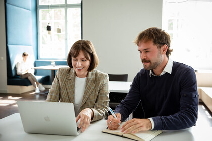 zwei Personen sitzen an einem Tisch und schauen gemeinsam auf einen aufgeklappten Laptop