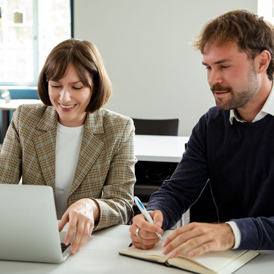 zwei Personen sitzen an einem Tisch und schauen gemeinsam auf einen aufgeklappten Laptop