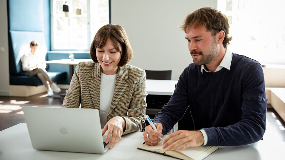 zwei Personen sitzen an einem Tisch und schauen gemeinsam auf einen aufgeklappten Laptop