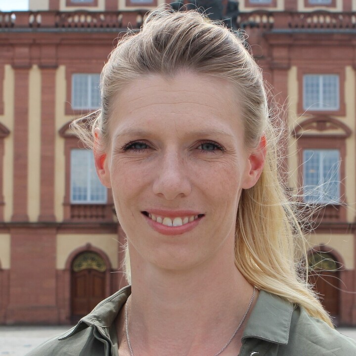 Eine Frau mit blonden Haaren und einem Zopf steht vor dem Schloss. Sie trägt eine dunkelgrüne Bluse.