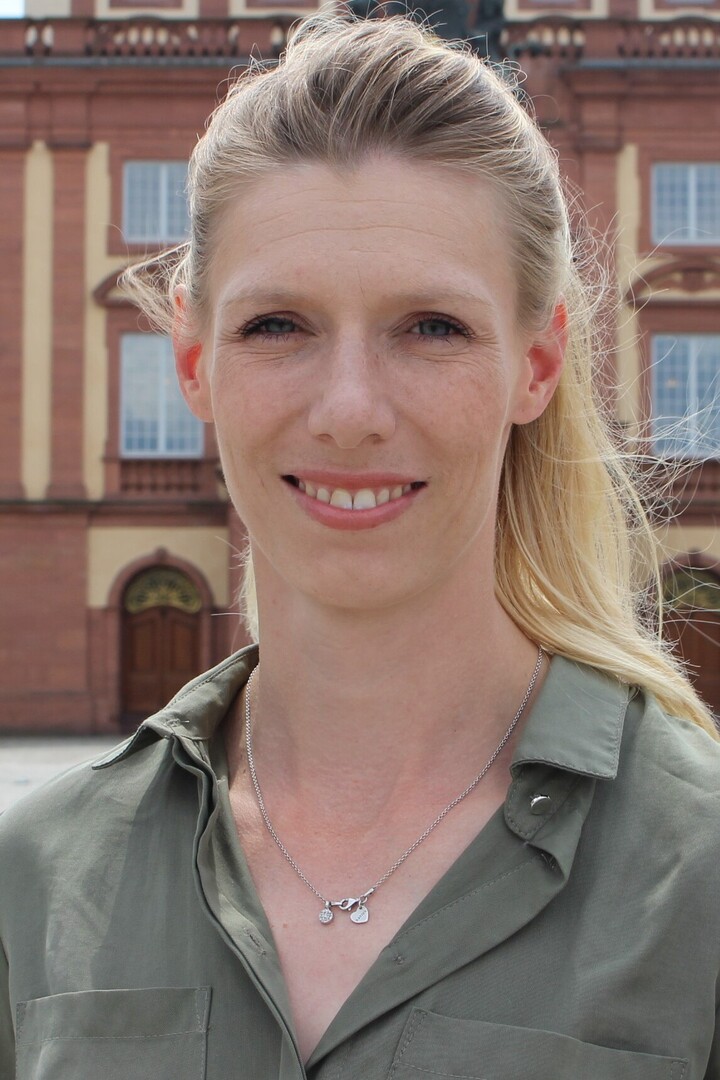 Eine Frau mit blonden Haaren und einem Zopf steht vor dem Schloss. Sie trägt eine dunkelgrüne Bluse.