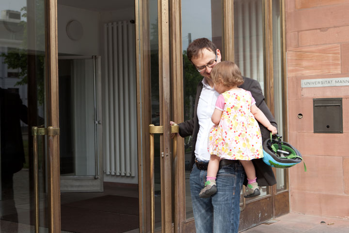 Ein Vater hält sein Kind auf dem Arm und lächelt. Gleichzeitig öffnet er eine Eingangstür zum Schloss Mannheim.