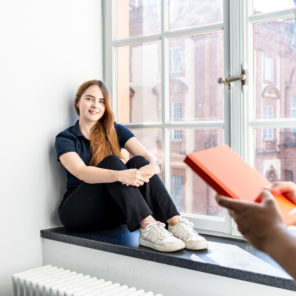 Eine Studentin sitzt an einem großen Fenster. Eine andere Studentin steht im Vordergrund und hält ein Buch in den Händen.