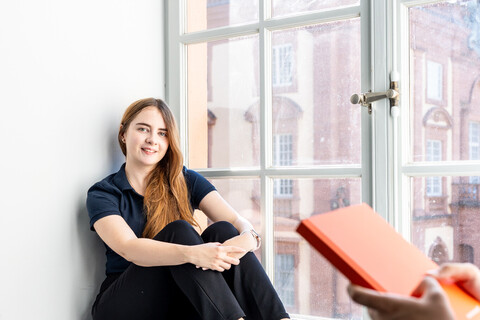 Eine Studentin sitzt an einem großen Fenster. Eine andere Studentin steht im Vordergrund und hält ein Buch in den Händen.