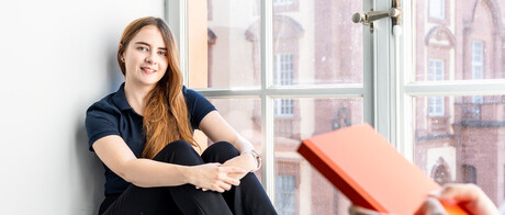 Eine Studentin sitzt an einem großen Fenster. Eine andere Studentin steht im Vordergrund und hält ein Buch in den Händen.