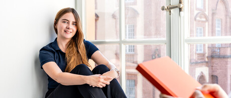 Eine Studentin sitzt an einem großen Fenster. Eine andere Studentin steht im Vordergrund und hält ein Buch in den Händen.