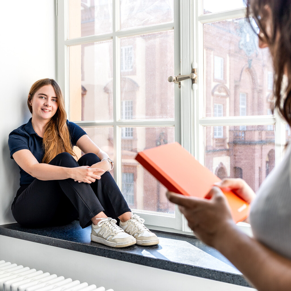 Eine Studentin sitzt an einem großen Fenster. Eine andere Studentin steht im Vordergrund und hält ein Buch in den Händen.