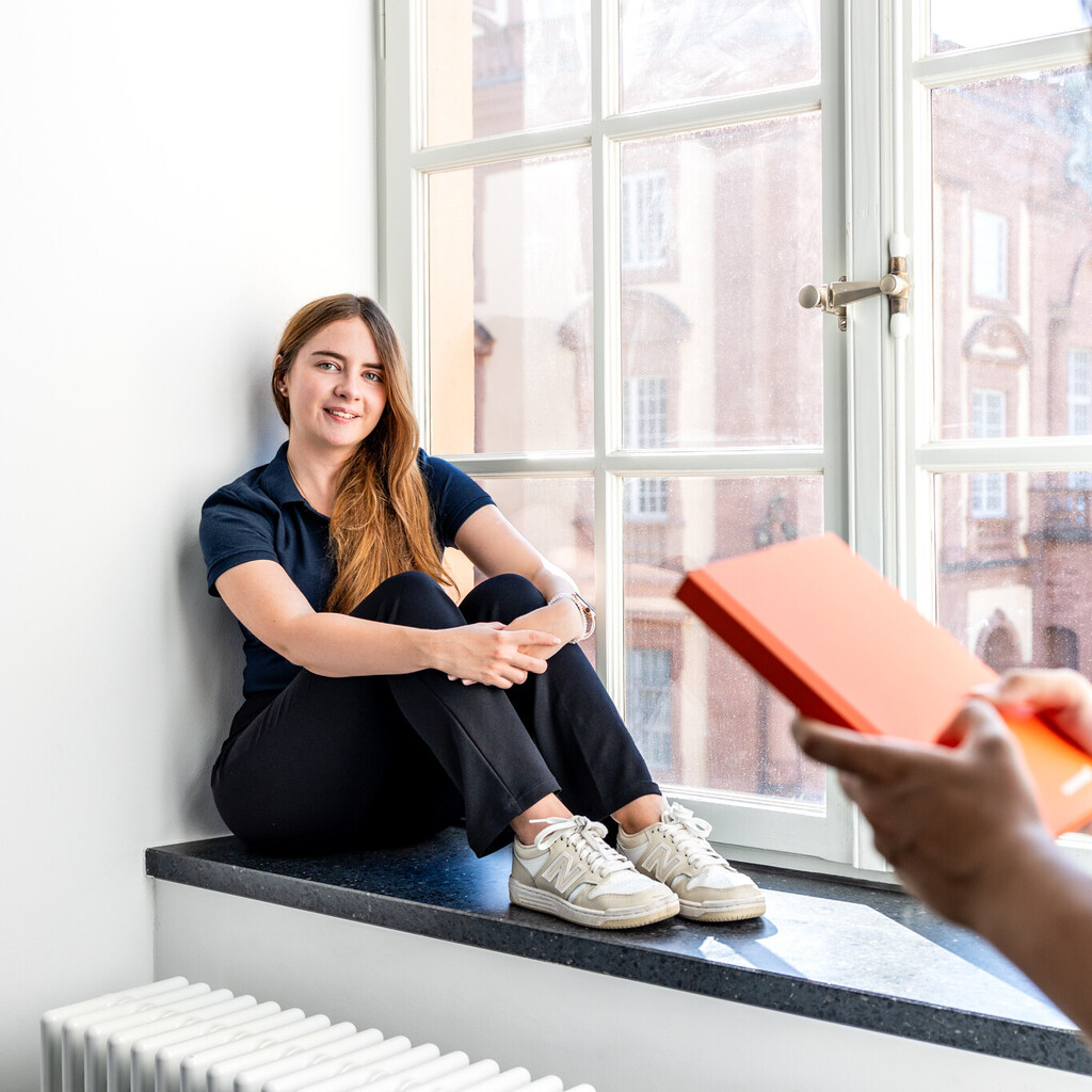 Eine Studentin sitzt an einem großen Fenster. Eine andere Studentin steht im Vordergrund und hält ein Buch in den Händen.