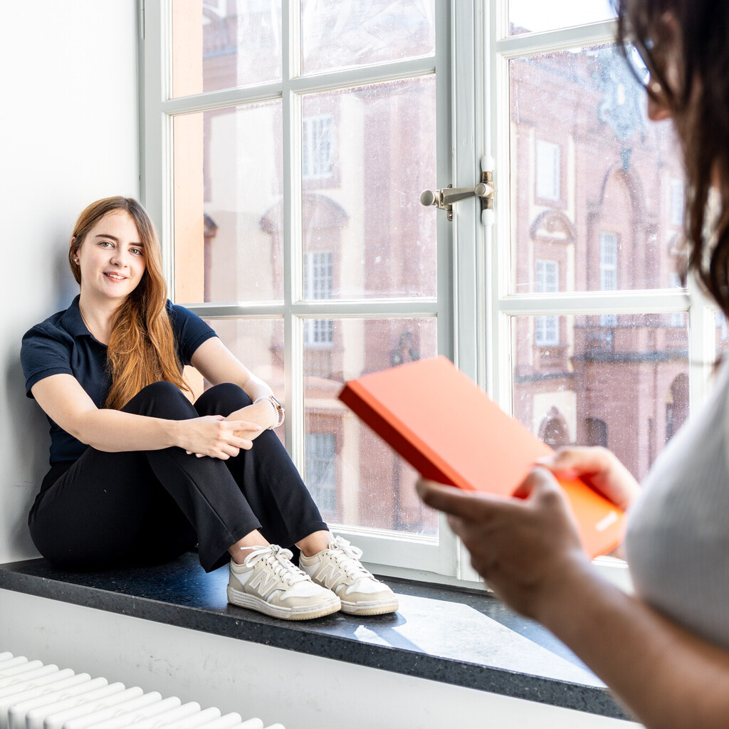 Eine Studentin sitzt an einem großen Fenster. Eine andere Studentin steht im Vordergrund und hält ein Buch in den Händen.