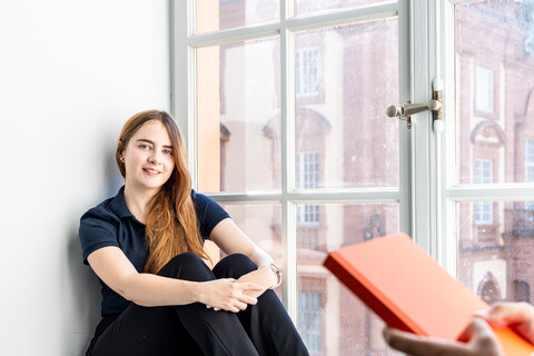 Eine Studentin sitzt an einem großen Fenster. Eine andere Studentin steht im Vordergrund und hält ein Buch in den Händen.