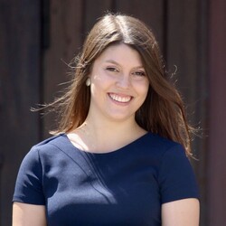A portrait of Annamaria Grossmann. She is smiling, has long brown hair, and is wearing a dark blue top. She is holding a book with the gold title “Masterarbeit” (Master’s Thesis).