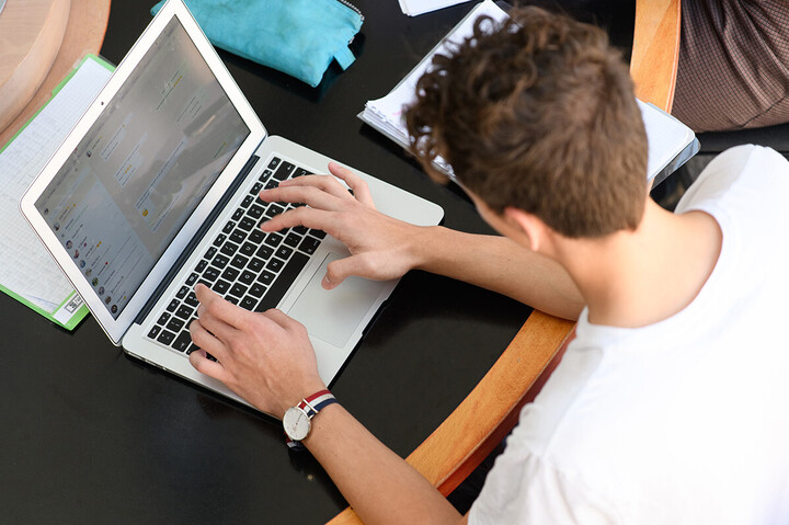A student sits at a table and writes on his laptop.