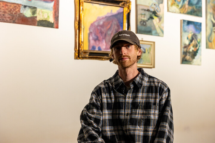 Der Künstler Jake Sandquist vor seinen ausgestellten Gemälden. Er schaut in die Kamera und trägt eine Kappe.