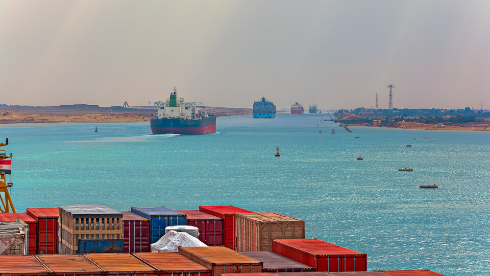 Im Vordergrund sieht man Container, im Hintergrund fahren Schiffe durch den Suez-Kanal