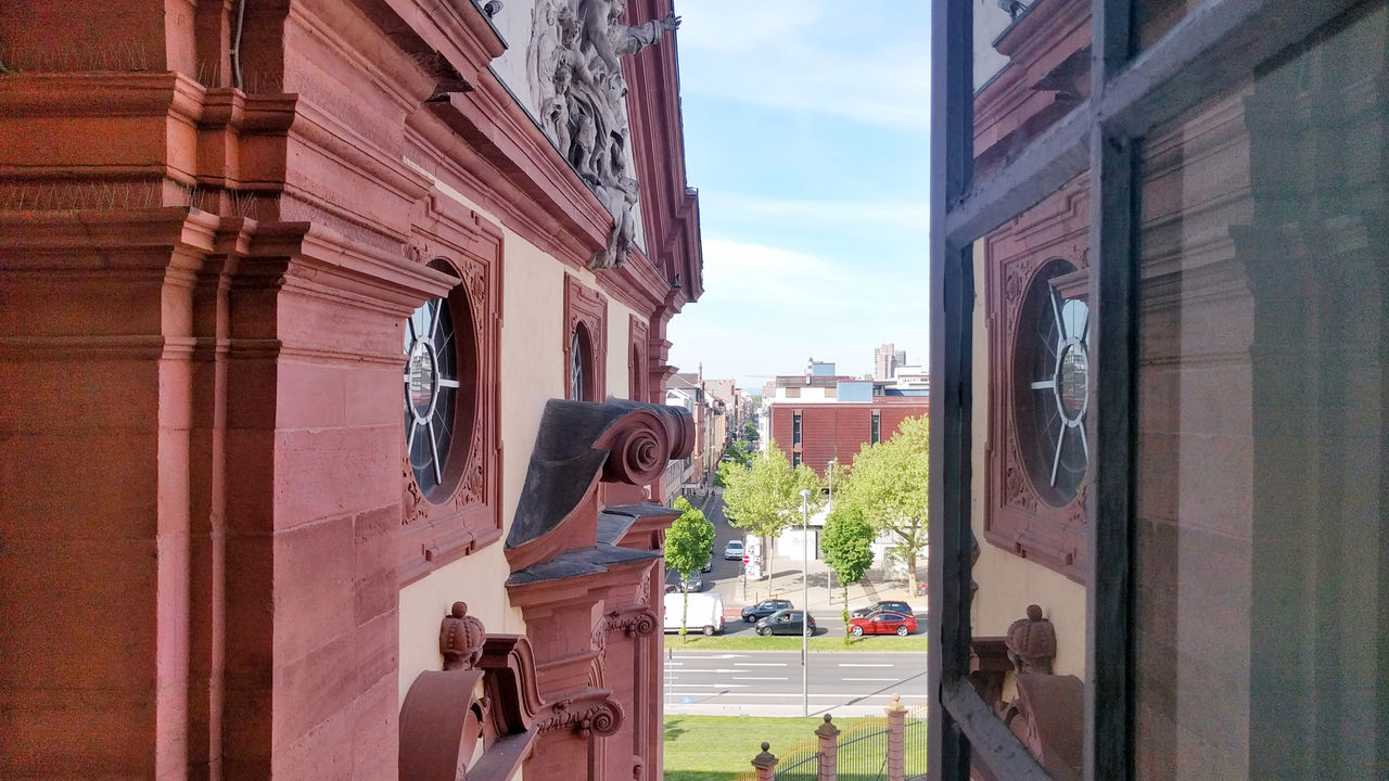Der Blick aus einem offenen Fenster des Mannheimer Schlosses. Die roten Mauern des Schlosses sind im Vordergrund. Im Hintergrund fahren Autos auf der Bismarckstraße direkt vor dem Schloss.
