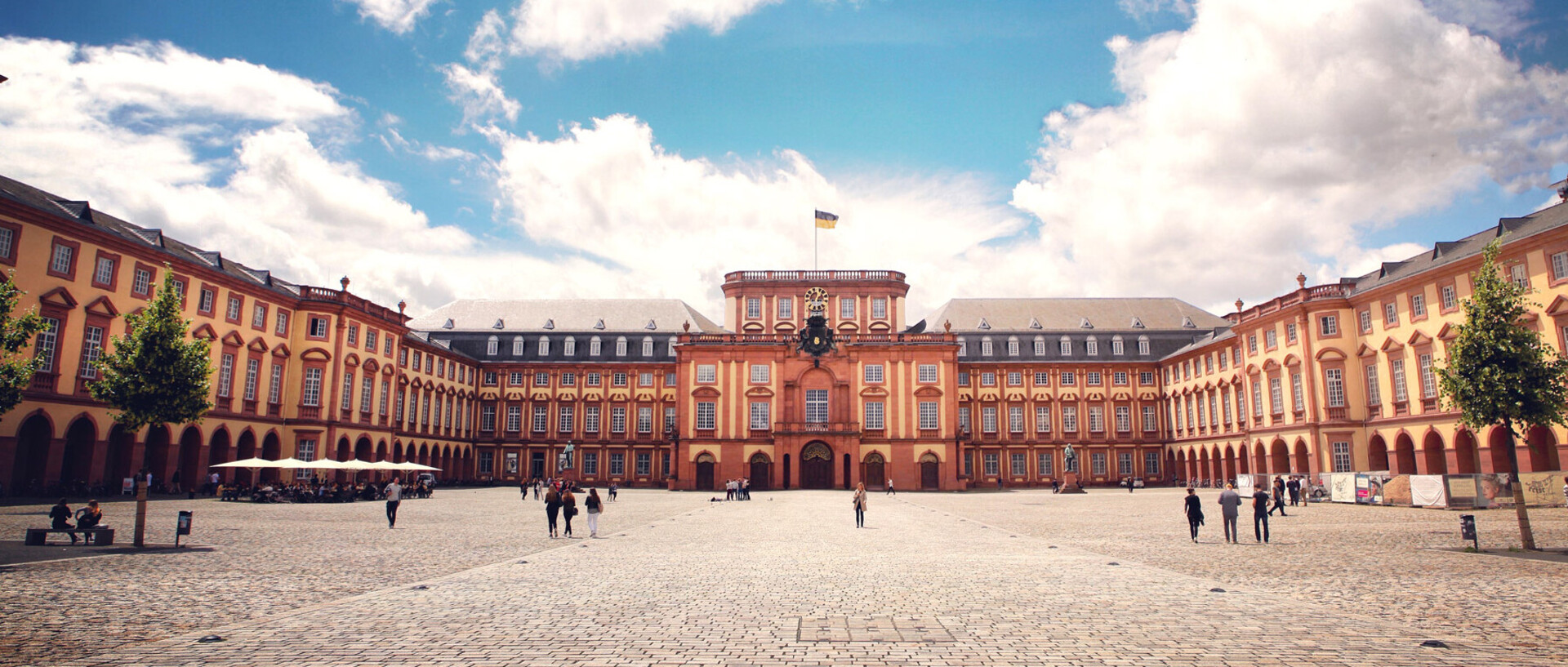 Das Barock-Schloss und der Ehrenhof der Universität Mannheim unter strahlend blauem Himmel. Das Schloss ist von unzähligen Fenstern, rotem Sandstein und einer gelben Fassade geprägt.