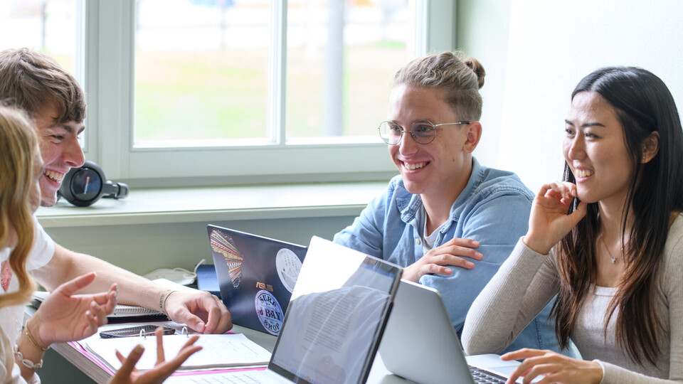 Vier Studierende sitzen vergnügt mit ihren Laptops an einem Fenster und arbeiten zusammen.