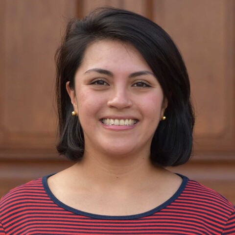 Eine lächelnde Person trägt ein rot-blau gestreiftes Oberteil undf steht vor einer braunen Holztüre. Die Person heißt Sofia Navarro-Báez.