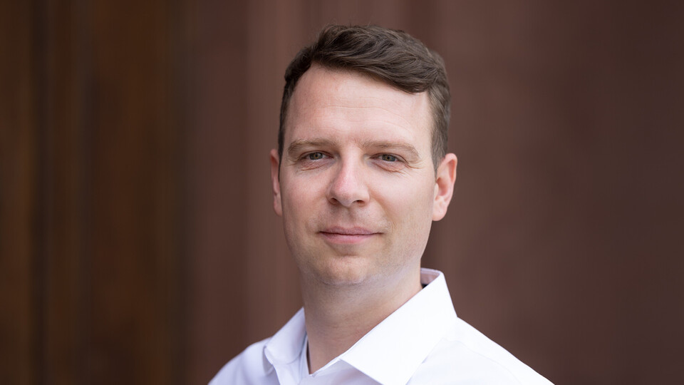 Oliver Pfäuti is wearing a white shirt in front of a brown background.