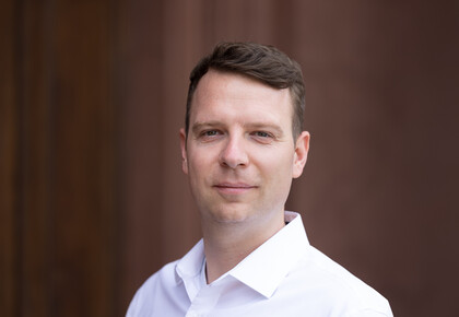 Oliver Pfäuti is wearing a white shirt in front of a brown background.