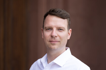 Oliver Pfäuti is wearing a white shirt in front of a brown background.
