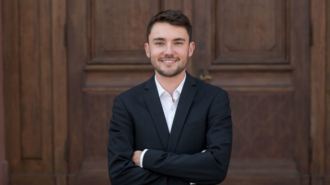 Daniel Klein. He is wearing a black suit and is standing in front of a brown door.