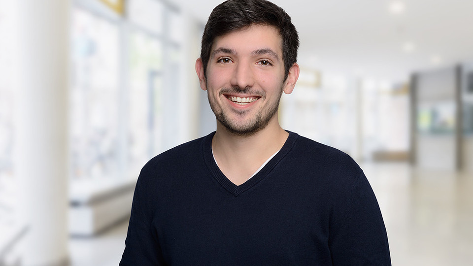 Portrait von Majed Dodin. Er trägt einen dunkelblauen Pullover und steht lächelnd vor einer Fensterfront.