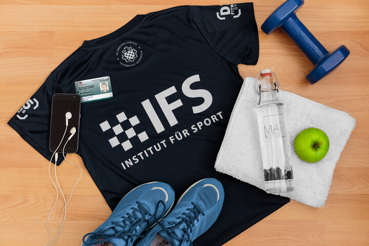 Eine Zusammenstellung von Merchartikeln des Instituts für Sport. Man sieht ein T-Shirt mit Logo, eine Wasserflasche, einen grünen Apfel, ein weißes Handtuch, blaue Sportschuhe und eine blaue Kurzhantel.