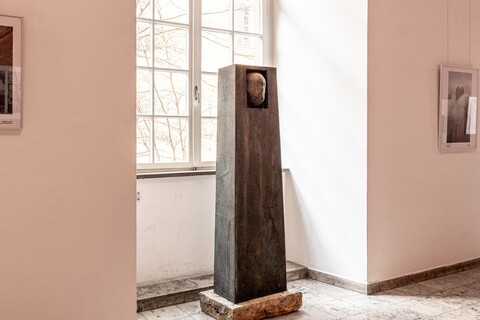 Bronzeplastik: Ein Gesicht ragt teilweise aus einem quaderförmige Block.
