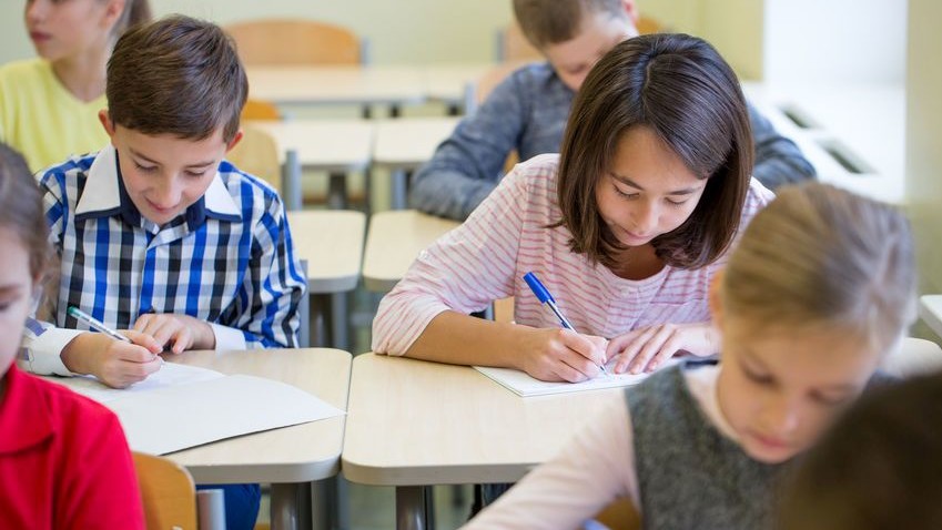 Schüler*innen sitzen nebeneinander an Tischen und schreiben auf ein Blatt Papier.