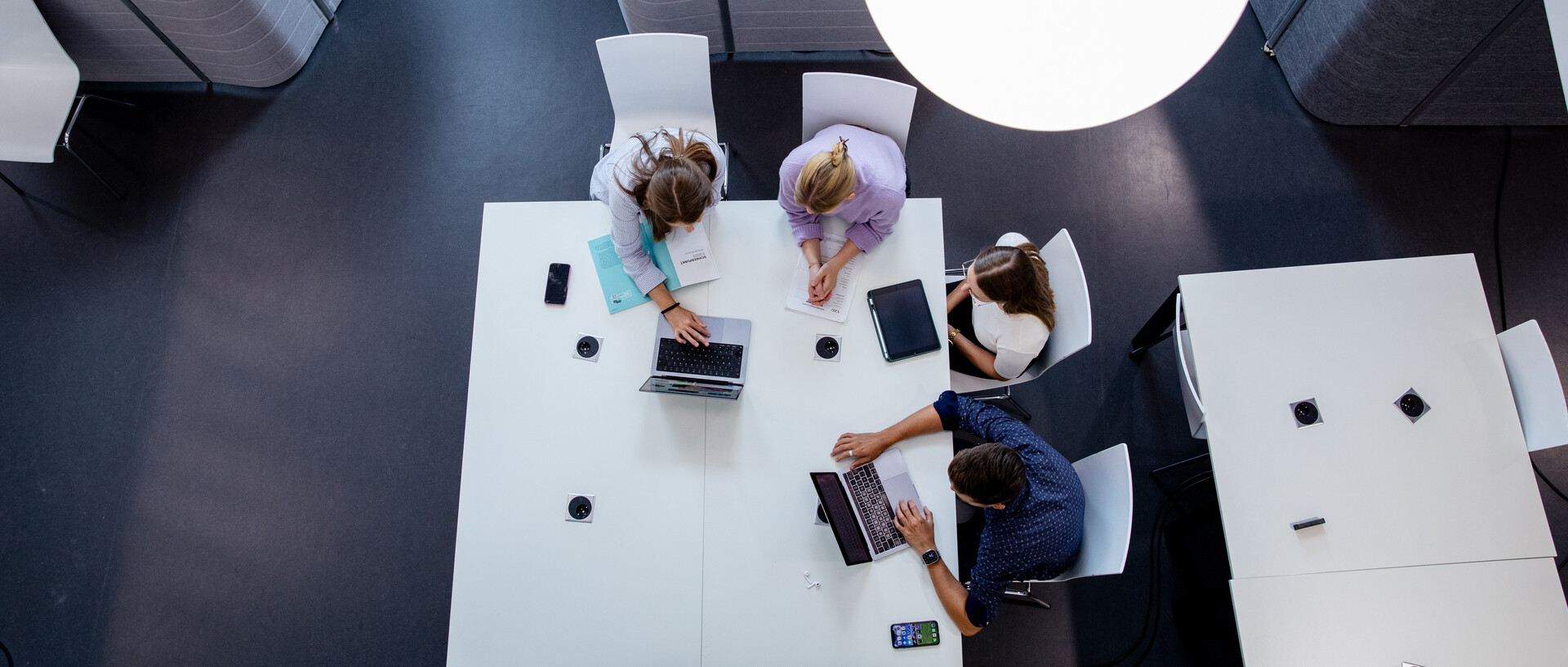 Vogelperspektive auf vier Personen bei einem Meeting an einem modernen, weißen Tisch in einem Büro mit dunklem Bodenbelag und einer großen Pendelleuchte über dem Tisch. Die Personen sind mit Laptops, Notizbüchern und Smartphones beschäftigt. 