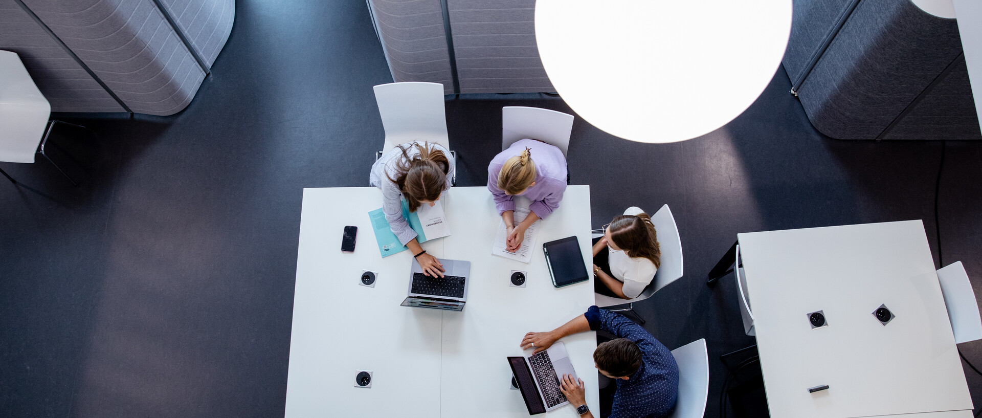 Vogelperspektive auf vier Personen bei einem Meeting an einem modernen, weißen Tisch in einem Büro mit dunklem Bodenbelag und einer großen Pendelleuchte über dem Tisch. Die Personen sind mit Laptops, Notizbüchern und Smartphones beschäftigt. 