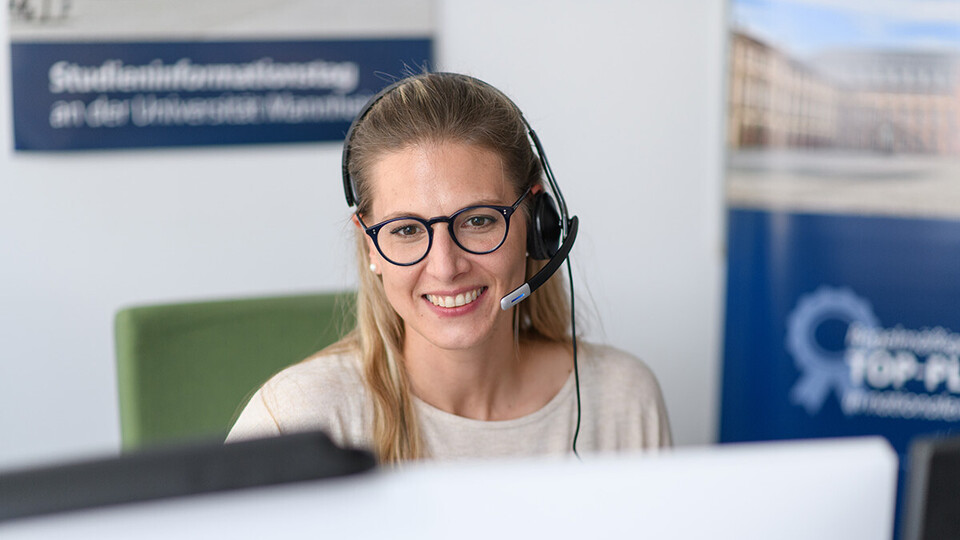 Eine Frau sitzt mit Kopfhörer in einem Büro, blickt in einen Bildschirm und lächelt.